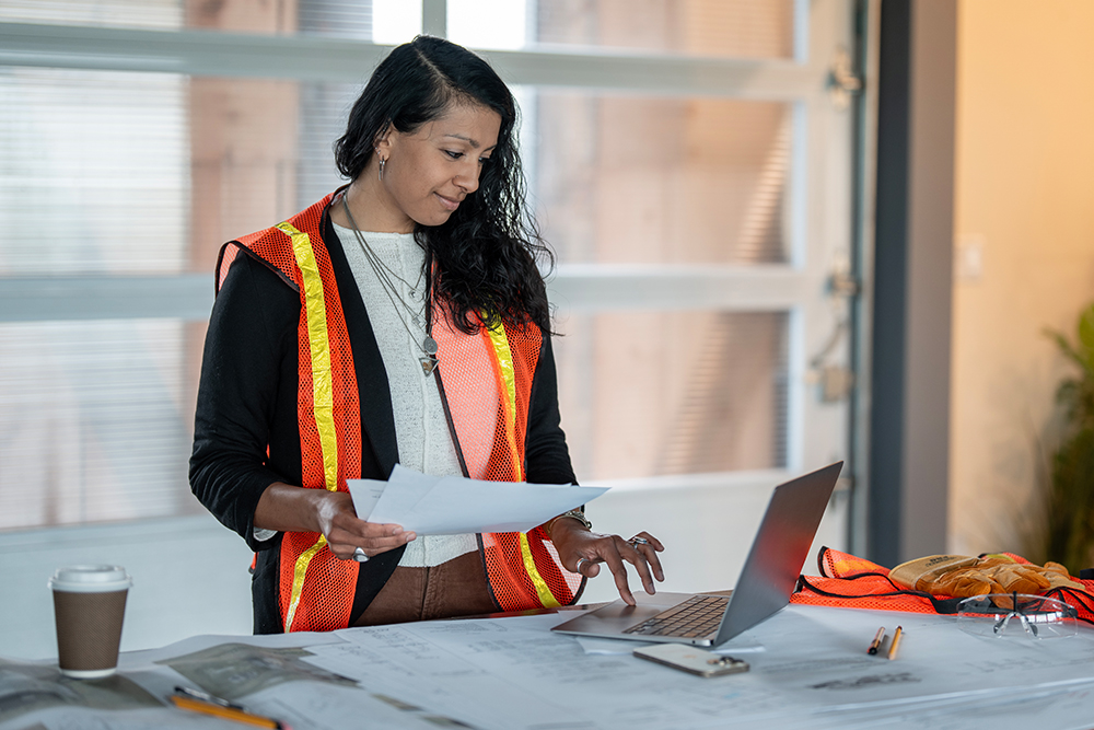 Civil engineer wearing an orange safety vest working on a computer at a construction site.