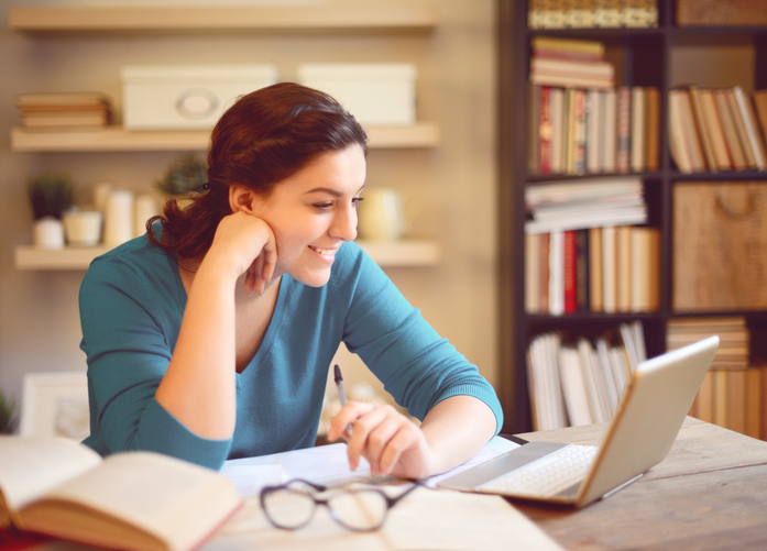 Female graduate student sitting at her home desk with her laptop, smiling as she studies with books open