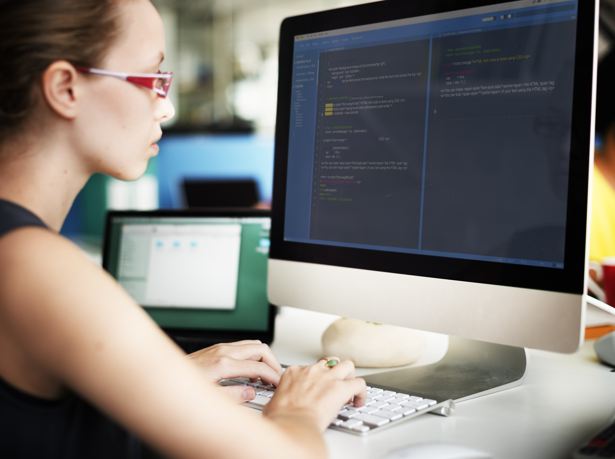 Female professional with red glasses sitting in front of a monitor, typing computer science coding 