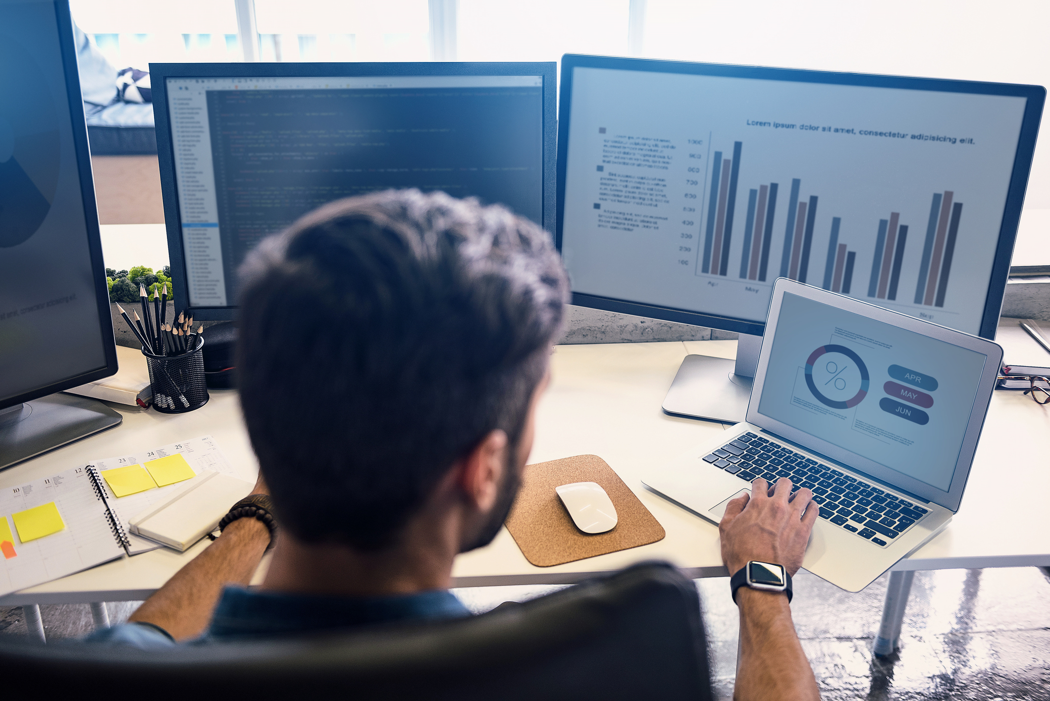 Male professional sitting at a desk with 3 computer monitors and a laptop, looking at analytics on each screen