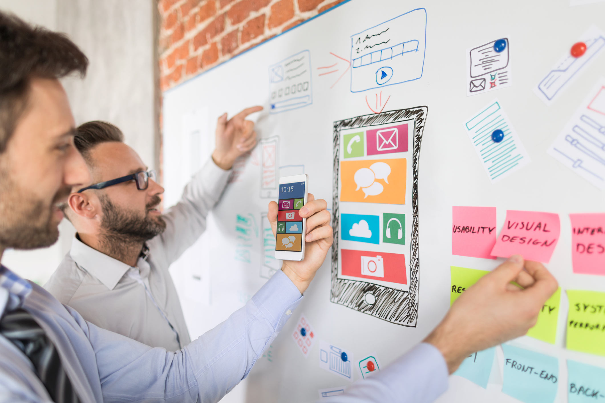 Two male professionals standing at a white board brainstorming ideas for social media design and web design. The white board has an image of an iPhone with colorful notes and ideas