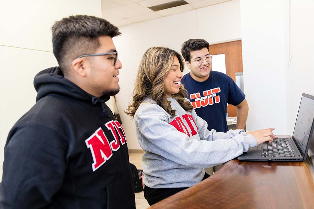 Three NJIT students smiling and reviewing information on a laptop.