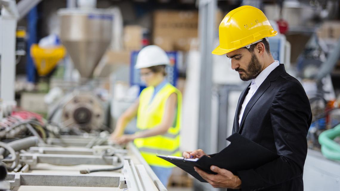 Professional engineer standing in a factory, wearing a hard hat, and taking notes