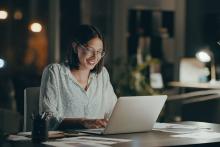 Online adult student smiles while typing on laptop in a home office setting.
