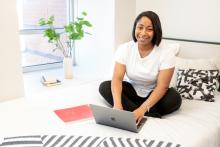 Black female adult learner sitting on her bed with her laptop doing school working smiling