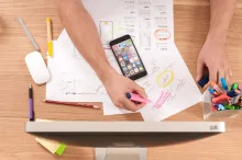 Image take from birdeye view of hands holding a highlighter and highlighting information on a design outline in front of a computer, mobile phone, and other design tools
