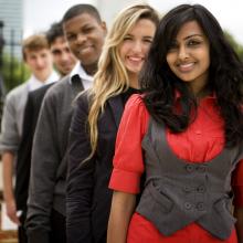 A group of diverse young professionals standing outside. The first is a young Indian lady wearing a bright red top and grey vest