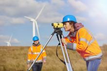 Female engineer wearing a hard hat and safety jacket, standing in front of windmills and using engineering equipment. Her male partner is standing behind her outside.